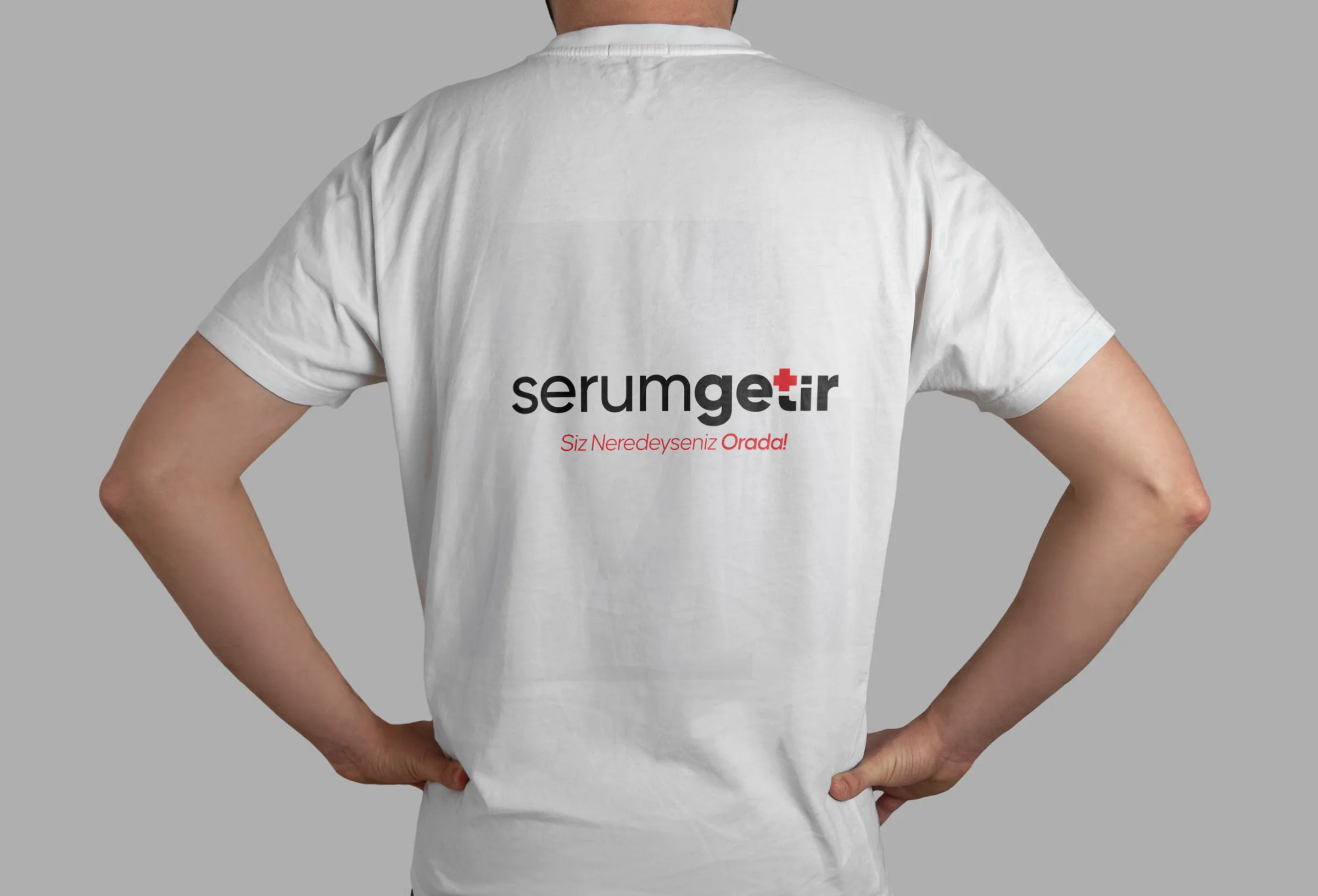 A person seen from behind, hands on hips, wearing a white T-shirt with the serumgetir logo and slogan across the back.